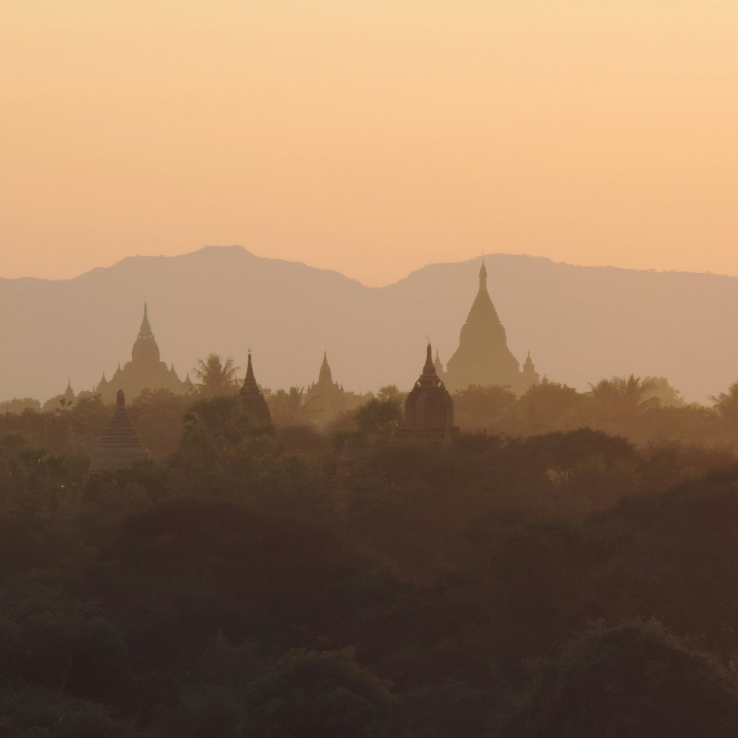 Silhouetted temples and trees at sunset with mountains in the background.