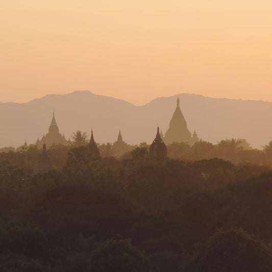 Silhouetted temples and trees at sunset with mountains in the background.