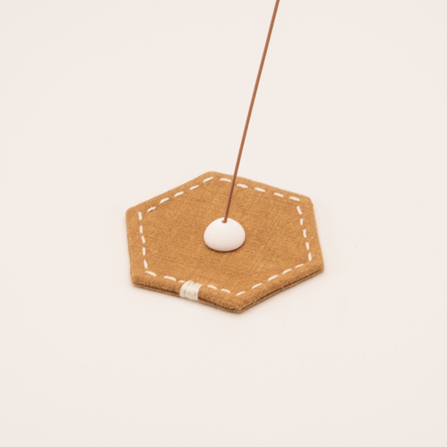 Minimalist incense stick holder with a white ceramic bead on a stitched hexagonal linen mat in warm tan color.