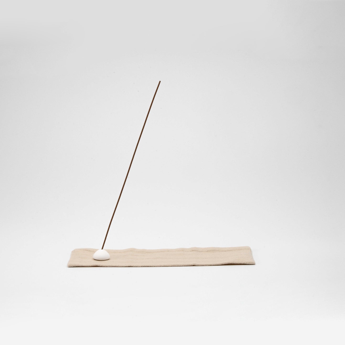 Minimal incense stick holder with a white ceramic bead on a beige striped linen mat, angled against a clean white background.