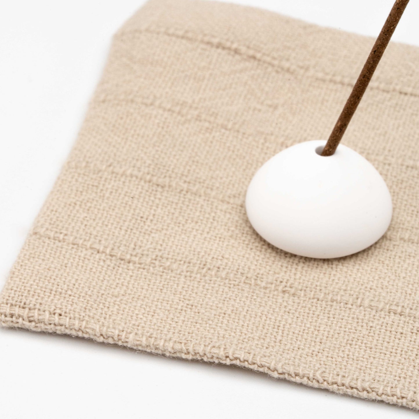 Close-up of textured beige linen mat with stitched stripes and a white ceramic incense holder supporting a lit incense stick.