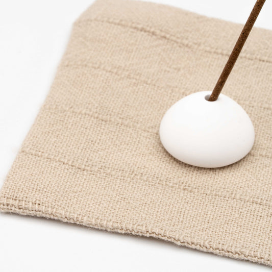 Close-up of textured beige linen mat with stitched stripes and a white ceramic incense holder supporting a lit incense stick.