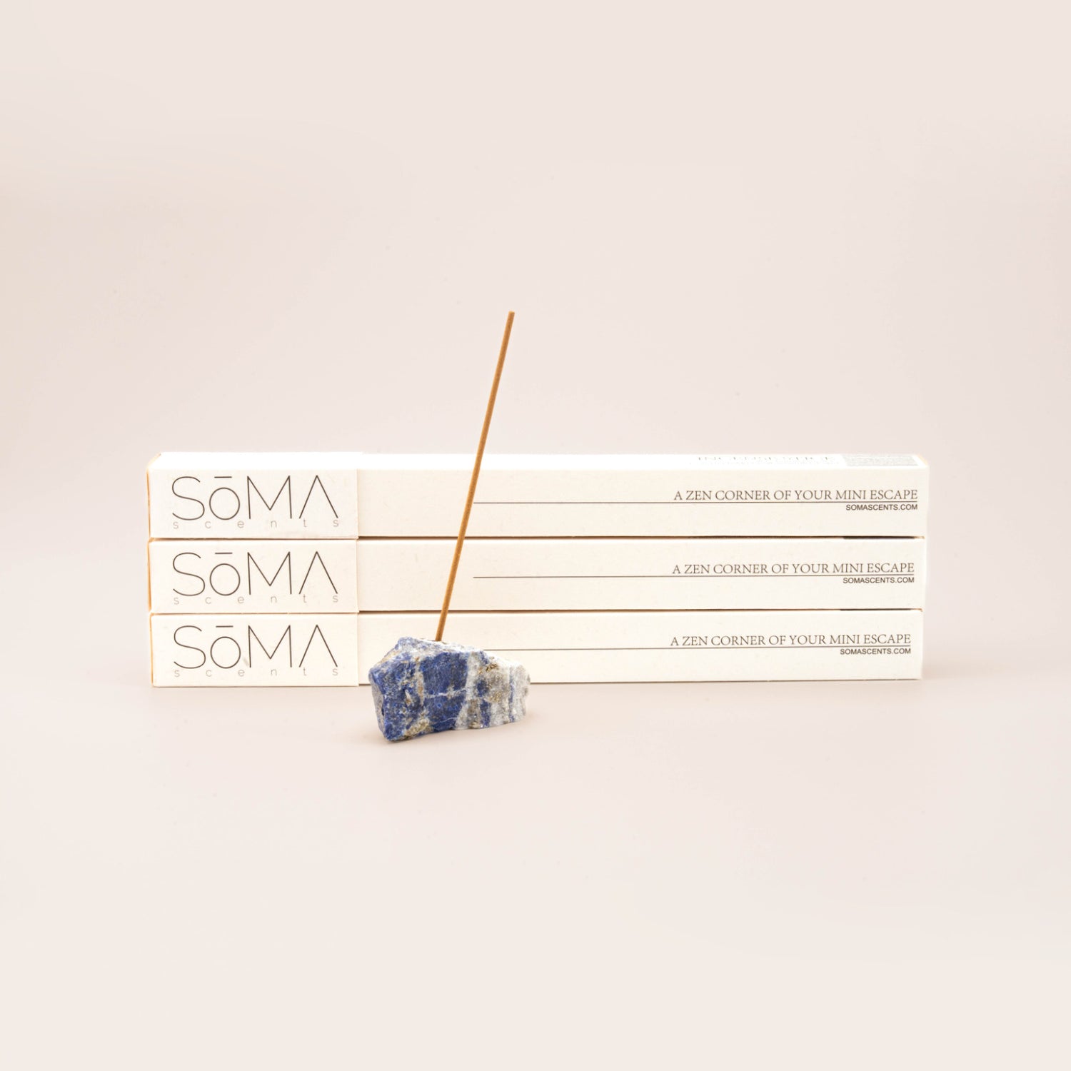 SomaScents incense stick burning in a blue sodalite gemstone holder, displayed in front of three stacked white incense boxes