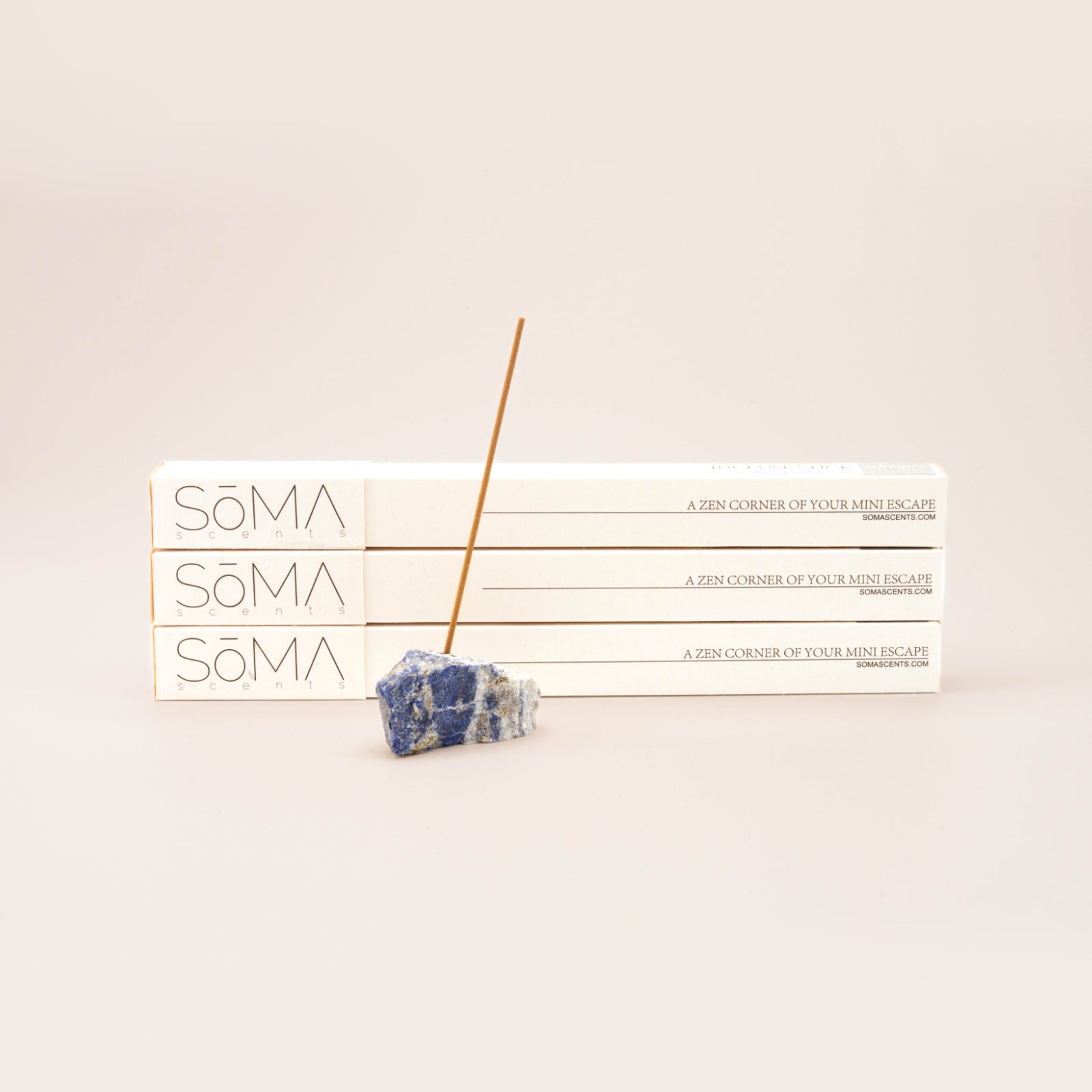 SomaScents incense stick burning in a blue sodalite gemstone holder, displayed in front of three stacked white incense boxes