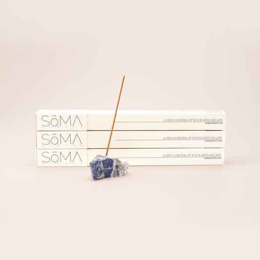 SomaScents incense stick burning in a blue sodalite gemstone holder, displayed in front of three stacked white incense boxes
