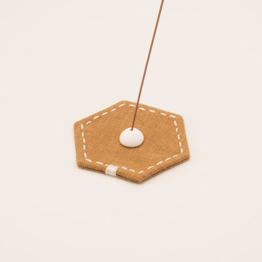 Minimalist incense stick holder with a white ceramic bead on a stitched hexagonal linen mat in warm tan color.