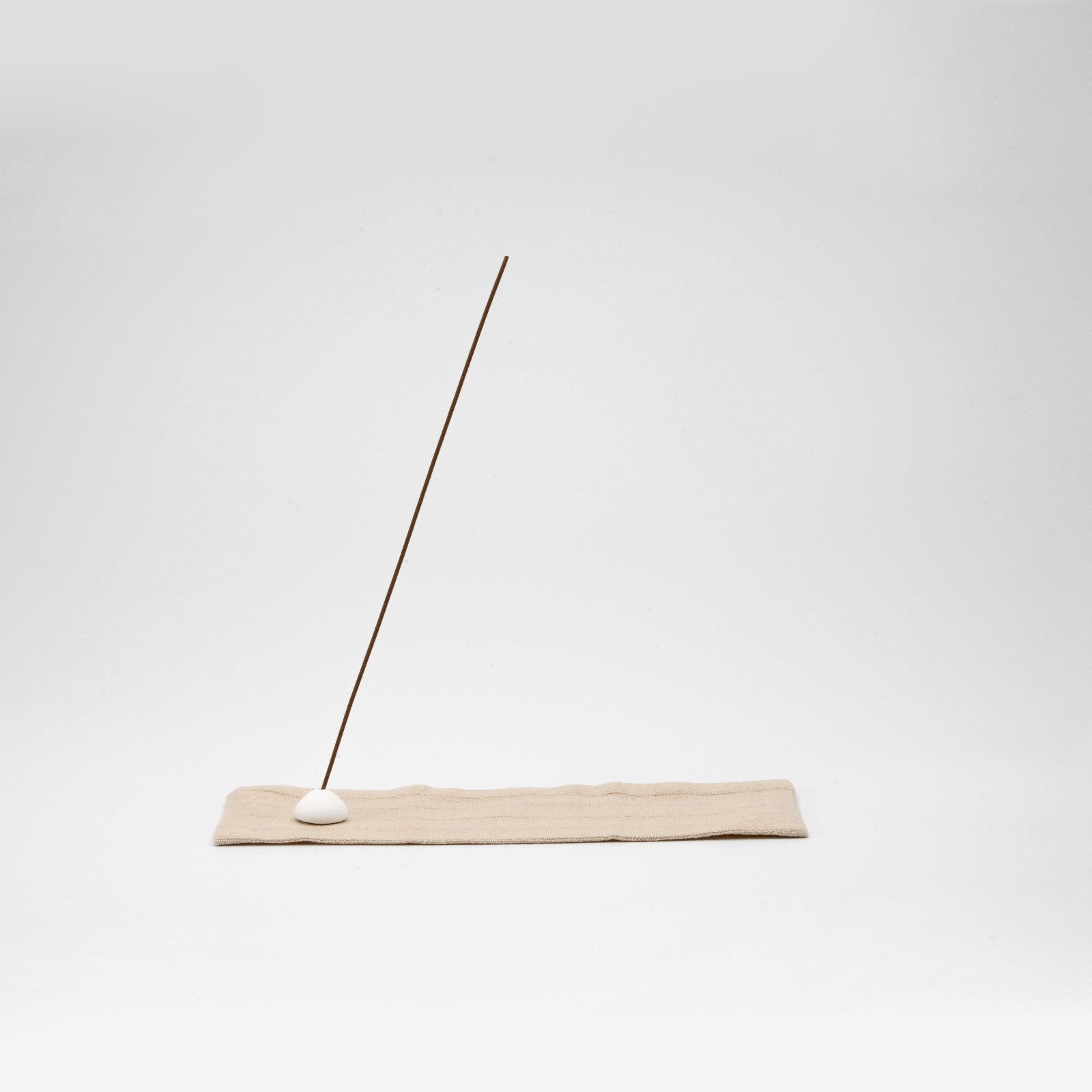 Minimal incense stick holder with a white ceramic bead on a beige striped linen mat, angled against a clean white background.