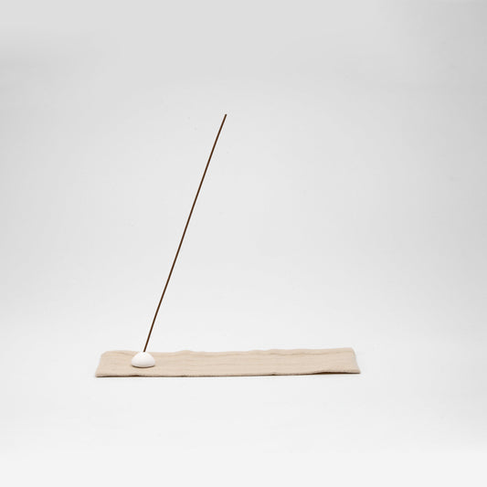 Minimal incense stick holder with a white ceramic bead on a beige striped linen mat, angled against a clean white background.